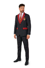 A man stands slightly to the right wearing the Harry Potter Suit Gryffindor™ by Suitmeister, which includes a black suit with red accents and an embroidered emblem on the chest pocket. He pairs it with a matching red and gold striped tie, a white shirt, and black shoes, set against a plain white background.