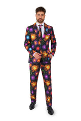 A man wearing Suitmeister's "Fireworks Black" NYE suit, featuring a vibrant fireworks pattern, stands against a white background. He complements the ensemble with a matching tie and white shirt, creating a festive and eye-catching look.