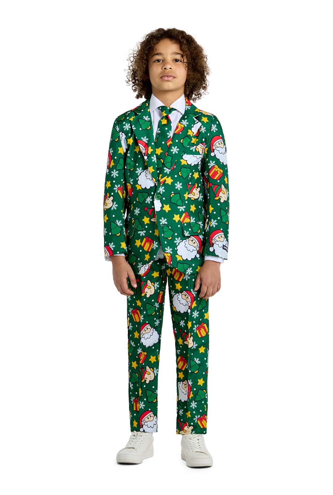 A child is dressed in the Santa Elves Green - Christmas suit by Suitmeister, which features a festive pattern adorned with Santa faces, stars, and gifts. The outfit is complete with a matching tie, and the child stands against a plain white background wearing white shoes.