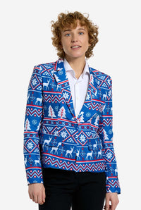 A person with curly hair is wearing the Suitmeister Christmas Jacket - Blue Nordic Jacket, which showcases a festive holiday pattern featuring snowflakes, trees, and reindeer. Underneath, they sport a white shirt and pair it with black pants, smiling slightly in a neutral pose.