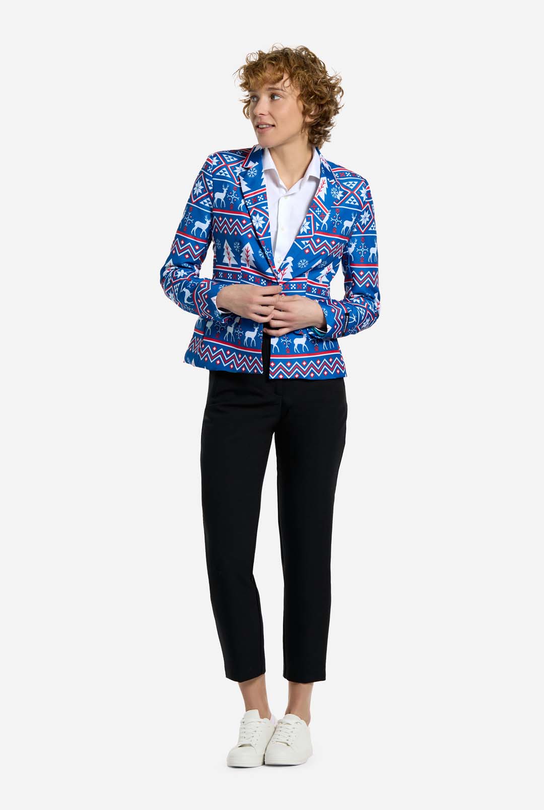 A person with curly hair wearing the Suitmeister Christmas Jacket - Blue Nordic Jacket, paired with a white shirt, black pants, and white sneakers stands against a plain background. They are looking to the side with hands partly clasped.