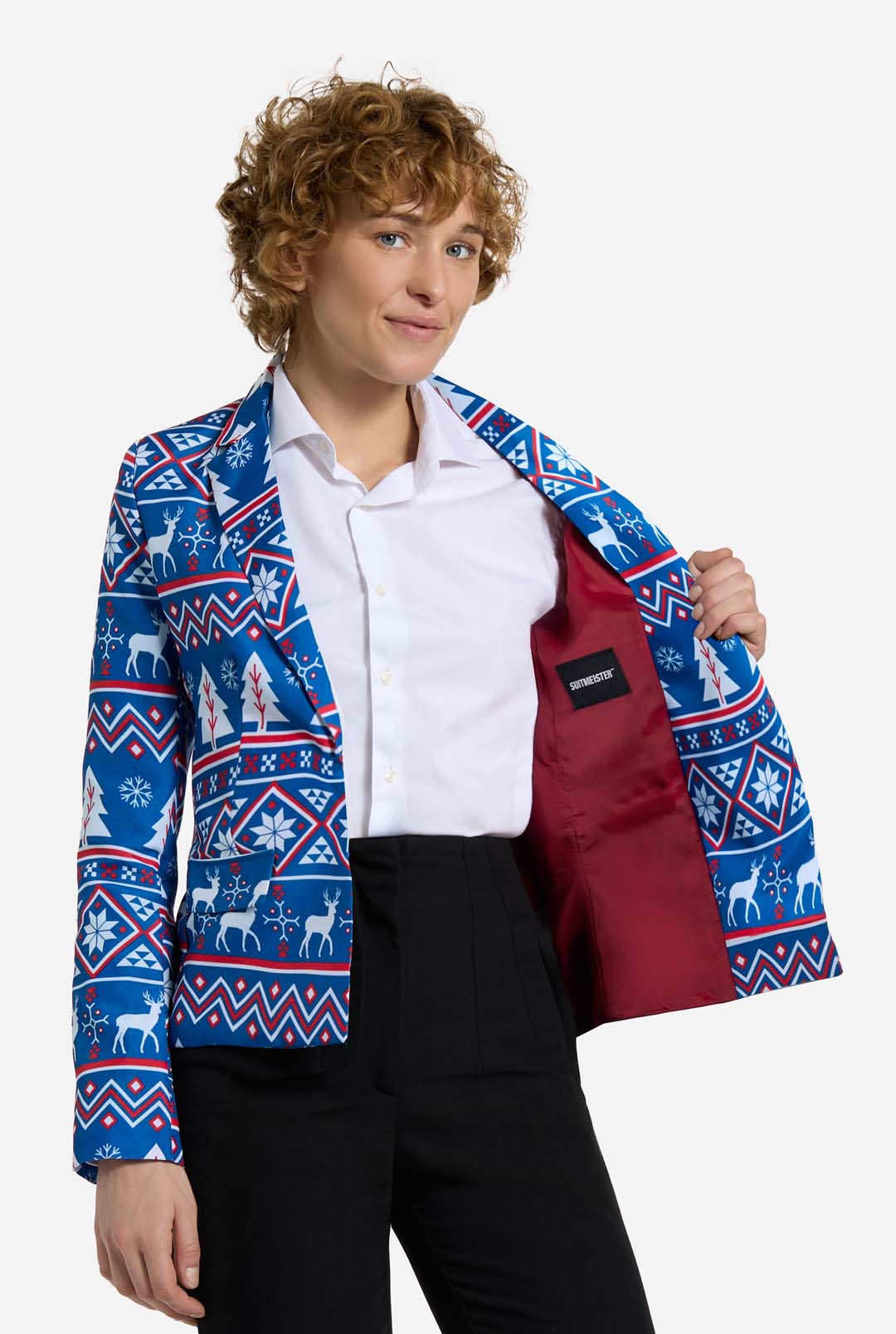 A person with curly hair models the Christmas Jacket - Blue Nordic Jacket by Suitmeister. This blue blazer showcases a festive winter pattern adorned with snowflakes and reindeer. They hold one side open to reveal a vibrant red lining, while wearing a crisp white shirt and black pants.