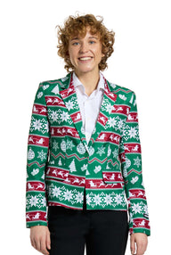 A person with curly hair is smiling while wearing the Suitmeister Christmas Green Nordic Jacket, a festive Christmas blazer featuring snowflakes, reindeer, and tree patterns in green, red, and white. They are standing in a white shirt and black pants.