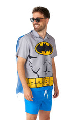 A person is showcasing the Suitmeister Batman™ - summer set, which includes a pair of sunglasses, a Batman-themed shirt featuring graphic muscles and a yellow utility belt, complemented by blue shorts. They are also wearing a blue cape that is attached to the shirt and smiling while looking to the side.