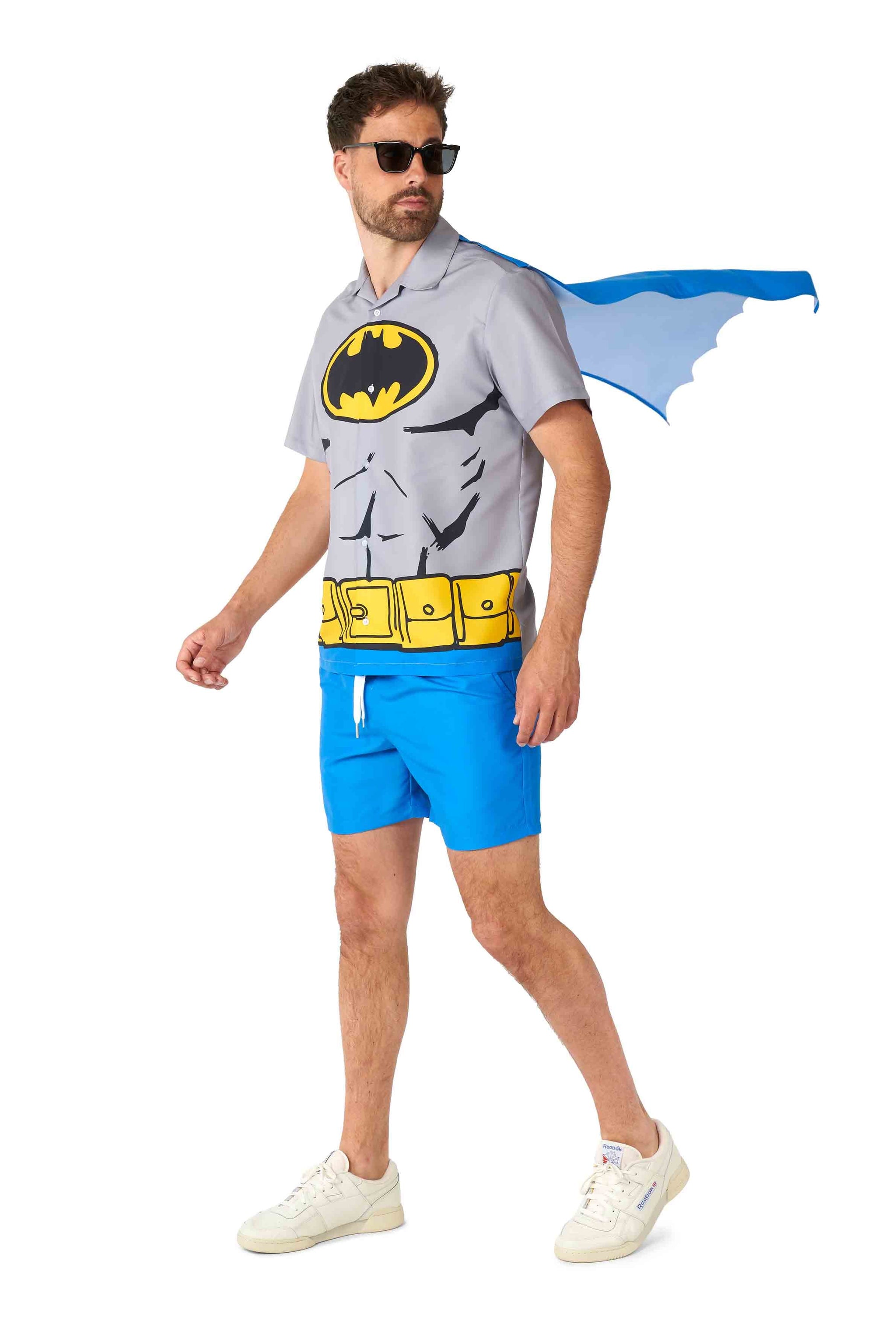 A man in sunglasses is sporting the Batman™ summer set by Suitmeister, featuring a gray and yellow superhero-themed shirt with a printed logo and a blue cape. He pairs this with blue shorts and white sneakers, exuding a playful vibe as he moves.