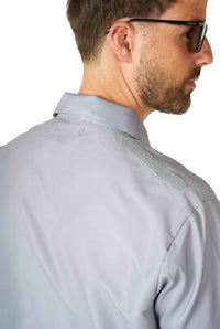 A man wearing sunglasses looks to the side, dressed in a light gray Batman™ - summer set from Suitmeister, featuring a reflective strip on the shoulder. The focus is on the back of the set and the reflective detail.