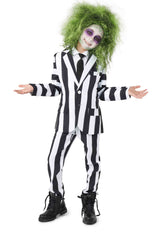 A child is dressed in a Suitmeister Beetlejuice suit paired with a tie and black boots, featuring green frizzy hair and a pale face accented by dark eyeshadow to create a spooky, ghostly appearance. With arms outstretched and palms facing up, the look is complete.