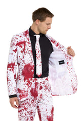 A man dressed in the Suitmeister Bloody Suit White, a Halloween-themed outfit featuring red splatter patterns, adjusts the open side of his jacket to reveal its lining. The suit is complete with a matching tie.