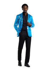 A man stands confidently wearing the Sequins Blue Blazer by Suitmeister, which shimmers brightly over a black shirt and pants. He poses with one hand in his pocket and the other adjusting the blazer against a plain white background.