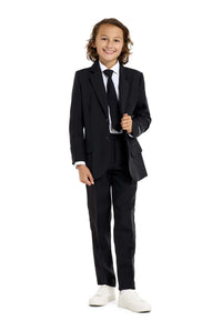 A young boy with shoulder-length hair confidently smiles in a Suitmeister Solid Black suit. He wears it with a white shirt, black tie, and white sneakers. Holding his tie, he stands against a plain white background.