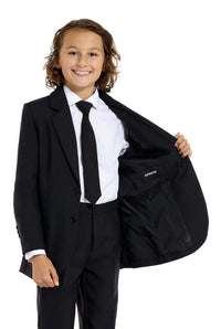 A smiling child with long hair wears the Suitmeister Solid Black suit—black jacket, white shirt, and black tie. The child is holding open one side of the jacket to reveal its lining against a plain white background.