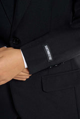 A person in a black Solid Black suit crosses their arms, revealing the Suitmeister embroidery on the cuff and a visible pocket on the jacket.