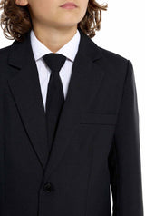 A young person wearing a Suitmeister Solid Black suit with a white shirt and black tie has long, curly hair and is posed against a plain white background.