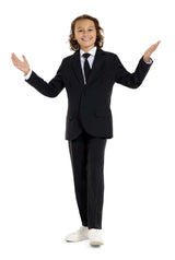 A smiling child with curly hair stands with arms wide open, wearing a Solid Black suit by Suitmeister accompanied by a white shirt, black tie, and white sneakers, all set against a plain white background.