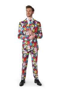 A man in a vibrant Suitmeister "Casino Icons" suit featuring graphic patterns of playing cards and dice stands confidently, with hands clasped and a slight smile. He completes the look with a white shirt, matching tie, and black shoes.