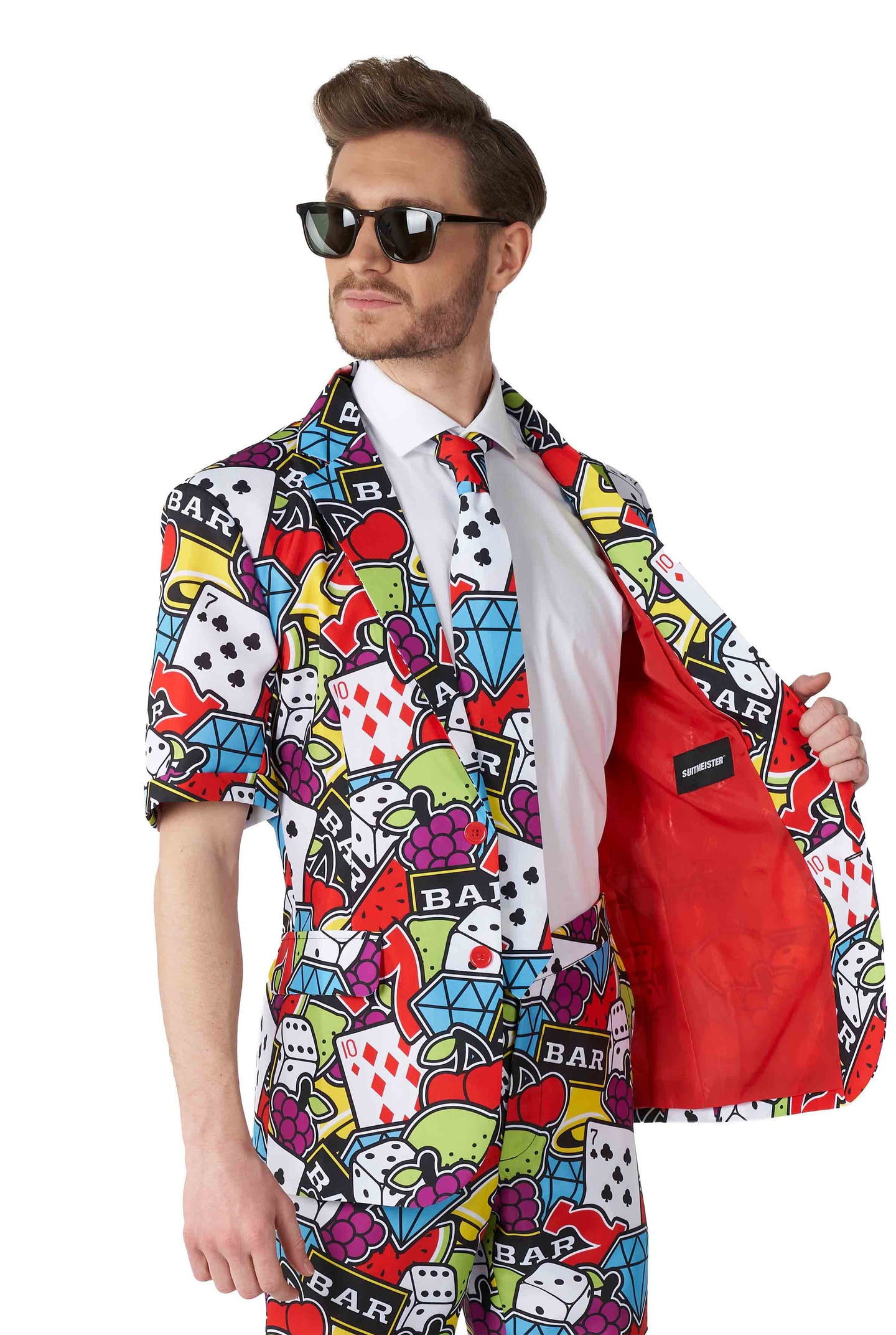 A man in sunglasses sports the vibrant Casino Icons - Carnival suit by Suitmeister, adorned with a lively casino-themed pattern showcasing dice, hearts, and slot machine icons. He opens the jacket to reveal its striking bright red lining while standing against a plain white background.