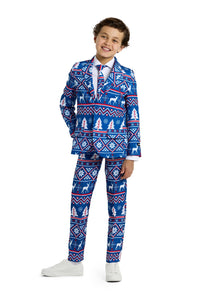 A smiling child stands with one hand in their pocket, wearing the Suitmeister Christmas Blue Nordic suit in festive blue, red, and white. The suit is adorned with holiday patterns including snowflakes, trees, and reindeer designs. They complete the look with white sneakers and a matching tie.