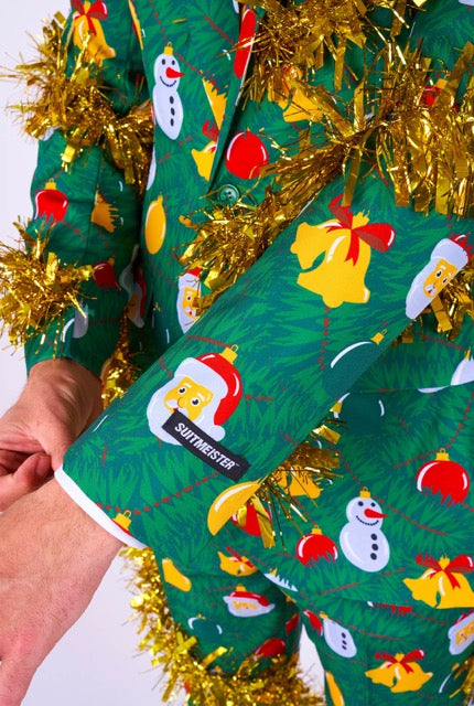 A person wearing a Christmas Deco Green suit covered in festive patterns featuring snowmen, bells, and ornaments. The suit is embellished with gold tinsel, and the sleeve sports a Suitmeister tag.