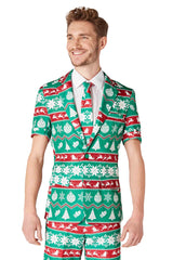 A man is dressed in a Suitmeister Christmas Green Nordic suit, showcasing a vibrant pattern of trees, snowflakes, and reindeer. He is smiling and gazing to the side, complemented by a matching tie and crisp white shirt.