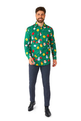 A person stands smiling, dressed in the "Christmas Ornaments Green" Christmas shirt by Suitmeister, featuring holiday-themed designs like snowmen and ornaments. They are also wearing dark blue pants and black shoes against a white backdrop.