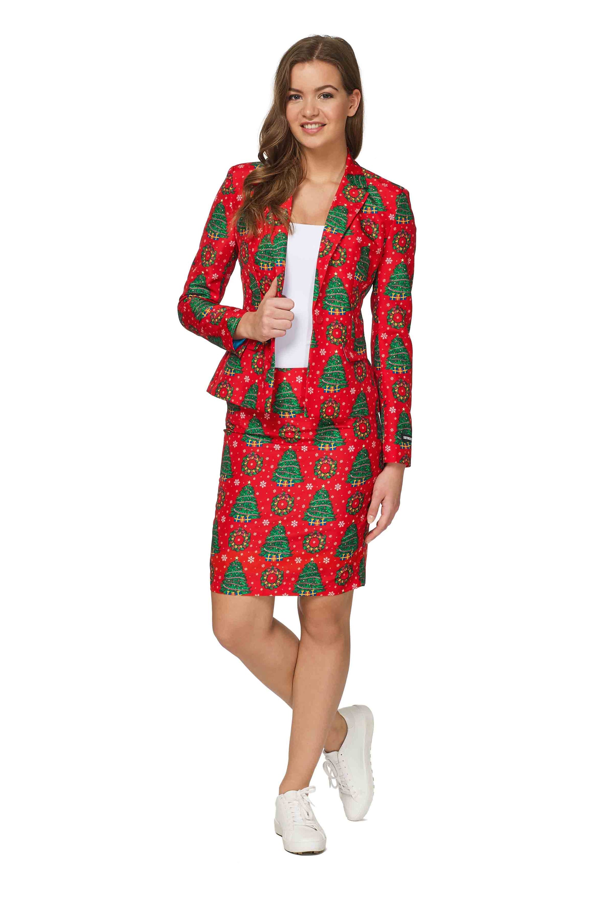 A person dressed in Suitmeister's Christmas trees - Christmas Suit, featuring a festive red skirt with a charming Christmas tree pattern, stands on one foot and grins. They have long hair and are also sporting white sneakers along with a white shirt beneath the blazer.