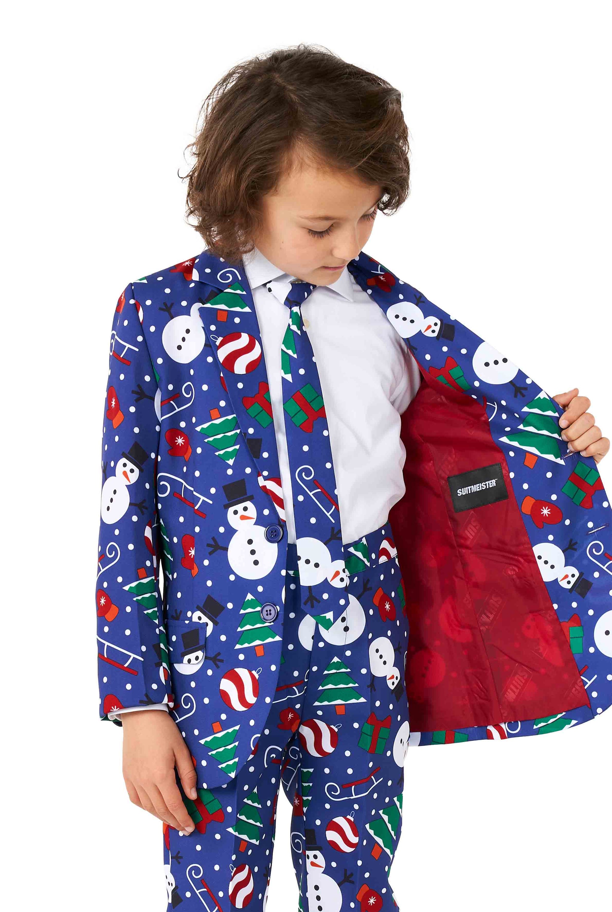 A child dressed in the Suitmeister Christmas Snowman Blue ensemble, featuring a blue suit adorned with snowmen, candy canes, trees, and gifts. The jacket is stylishly open to show off its vibrant red lining while the child sports a matching tie and gazes downward to the side.