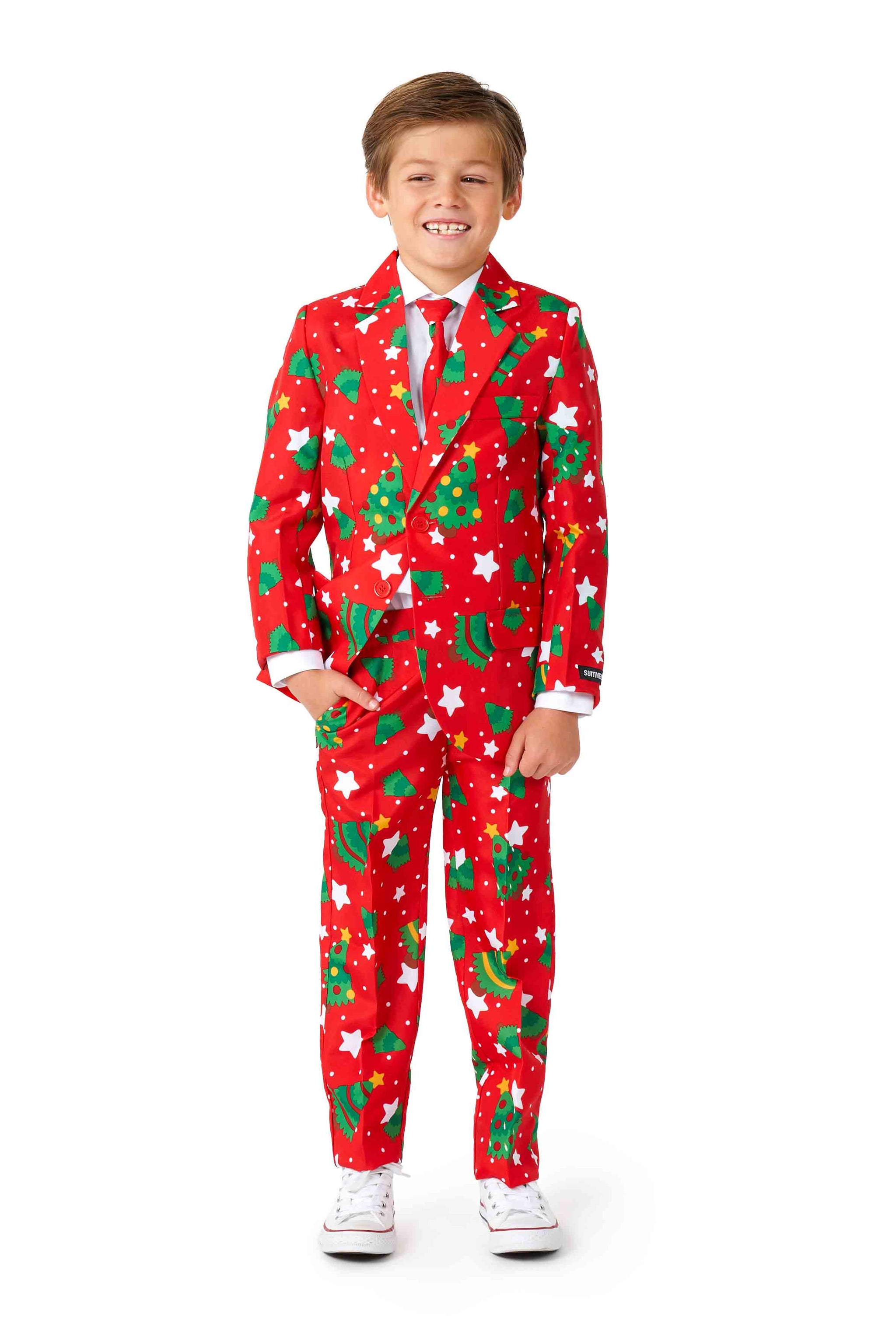 A young boy beams in a Suitmeister Christmas Trees Stars Red suit, adding festive cheer with its vibrant red hue and decorations of Christmas trees, stars, and candy canes. He pairs it with a white shirt, a red tie, and white sneakers while standing against a simple white background.