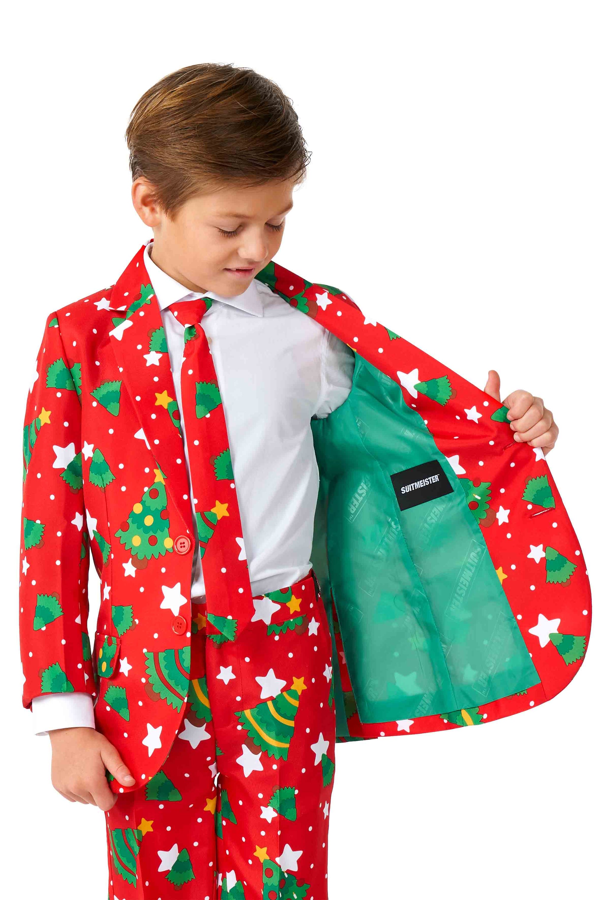 A young boy is dressed in a Suitmeister Christmas suit called "Christmas Trees Stars Red," featuring a festive red color adorned with green Christmas trees, stars, and various holiday designs. He is revealing the green interior lining of the jacket, which includes a label. Underneath, he is wearing a white shirt paired with a coordinating tie.