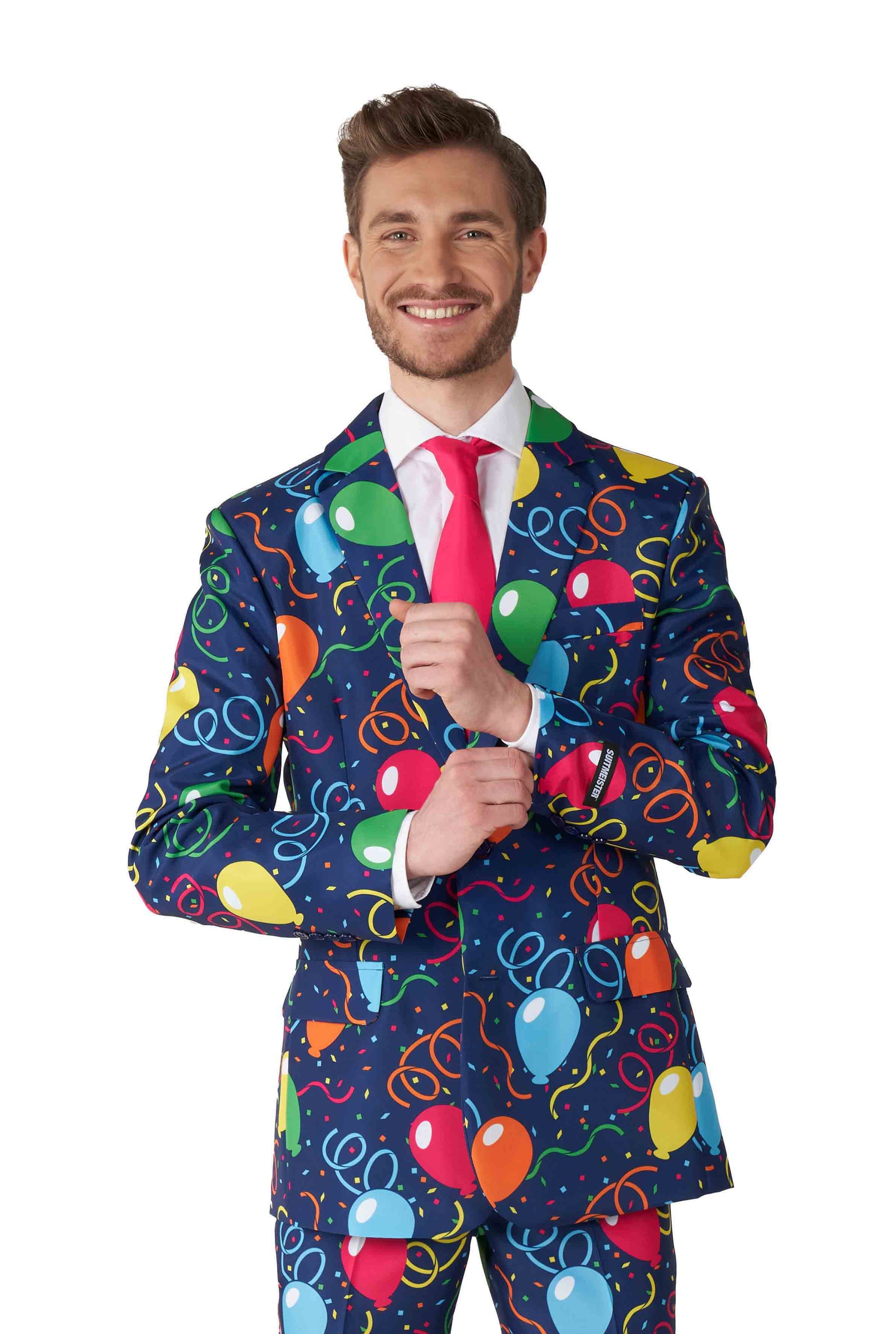 A person smiling, dressed in the Suitmeister "Confetti Balloons Navy" suit, featuring vibrant balloon and streamer patterns on a dark blue background. The outfit is complemented with a pink tie and white shirt, as the individual adjusts their shirt cuff.