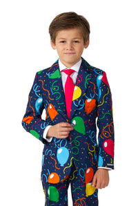 A young boy dressed in a vibrant Confetti Balloons Navy suit by Suitmeister, featuring festive balloon and confetti patterns. The ensemble is complemented with a white shirt and red tie as he stands facing the camera, smiling slightly.