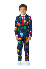 A young boy stands wearing the Suitmeister Confetti Balloons Navy suit, paired with a bright pink tie and white sneakers. The plain white background highlights his vibrant outfit.