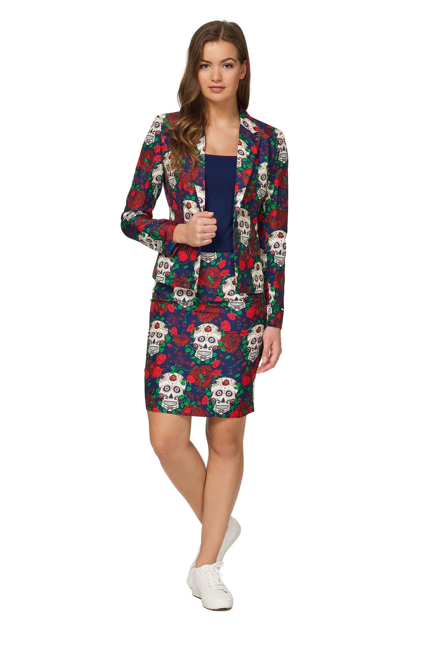 A woman stands wearing the Suitmeister "Day of the Dead - Halloween outfit," featuring a colorful blazer and skirt adorned with skulls and floral patterns. She pairs it with a navy top and white sneakers, set against a plain white background.