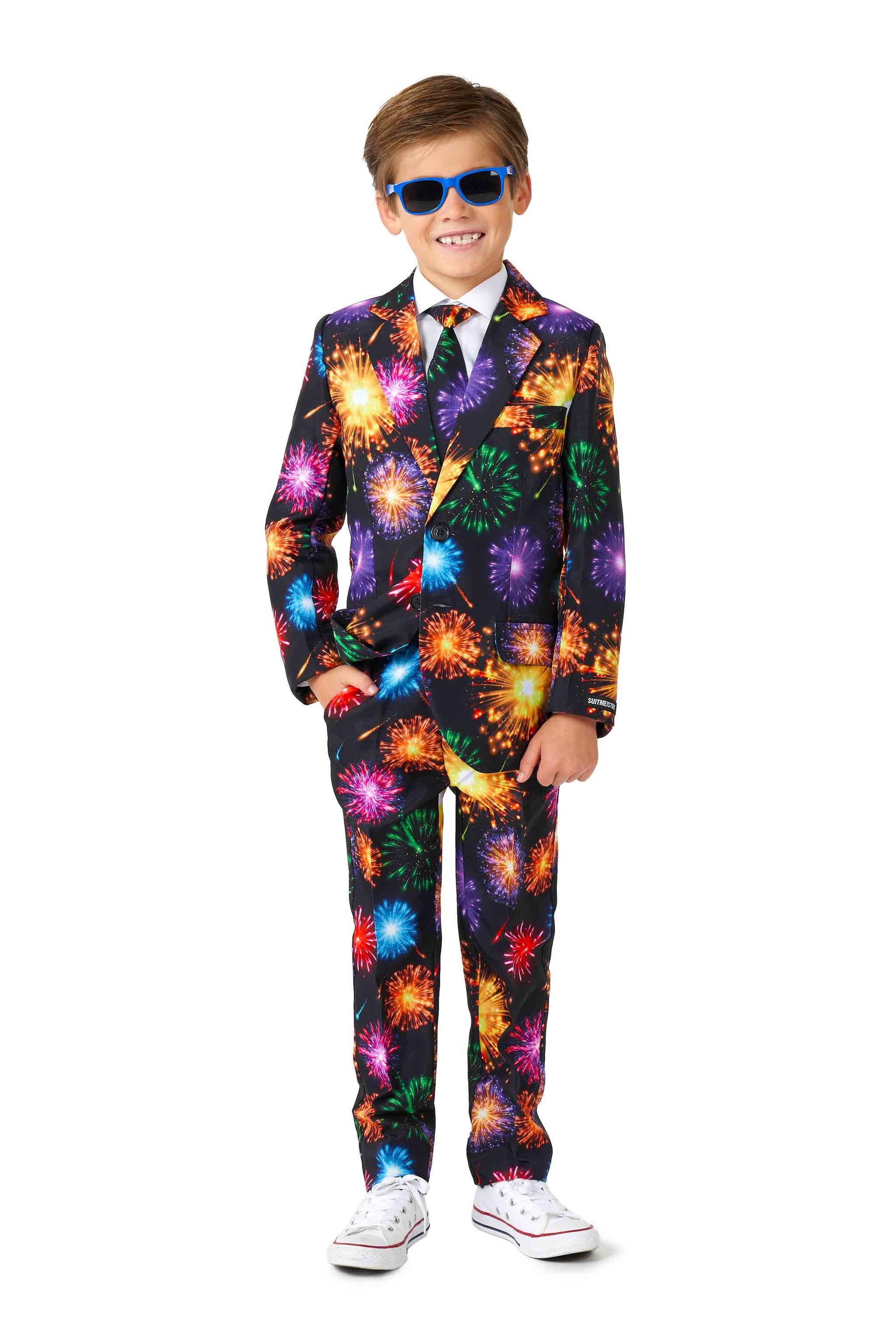 A child dressed in the Suitmeister's Fireworks Black ensemble, featuring a vibrant fireworks pattern, blue sunglasses, and white sneakers. The suit includes a matching tie, and the child is standing with one hand in their pocket, smiling.