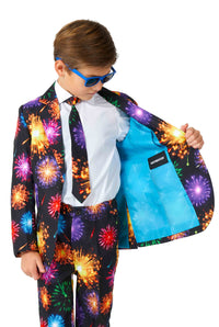 A young boy is dressed in Suitmeister's Fireworks Black NYE suit, featuring a vibrant fireworks pattern. He's sporting blue sunglasses and reveals the bright blue lining of the suit, where a label can be seen. The ensemble is completed with a white shirt and matching tie.