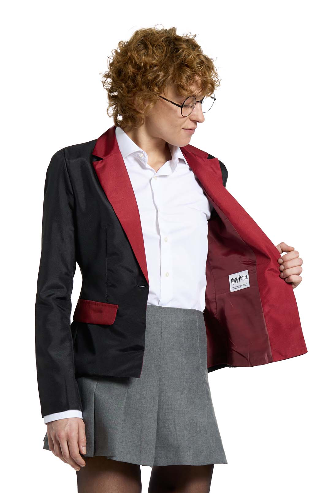 A person with curly hair and glasses is dressed in a Suitmeister Harry Potter Gryffindor™ Blazer, detailed with a red lining, paired with a white shirt and grey skirt. They are holding open one side of the blazer to show the label inside.