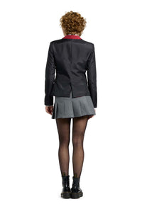 A person with curly hair, dressed in a Suitmeister Harry Potter Gryffindor™ Blazer, paired with a gray pleated skirt, sheer black tights, and black boots, is standing facing away from the camera against a plain white background.