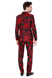 A man stands facing away from the camera, dressed in the Suitmeister Halloween Black Blood outfit, featuring a striking red abstract pattern on a black suit. His dark hair is neatly styled, and he complements the look with black shoes against a plain white background.