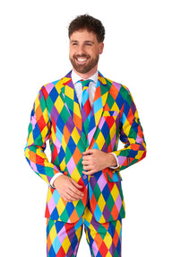 A smiling man is dressed in the Suitmeister's Harleclown - Mardi Gras suit, which comes with a matching tie. The suit boasts a vivid diamond pattern and a striking combination of bold colors such as red, blue, yellow, and green. The backdrop is plain white.