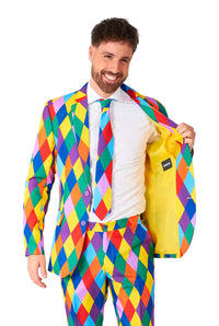 A man is smiling while wearing a vibrant Harleclown Mardi Gras suit by Suitmeister, featuring a colorful diamond pattern with a matching tie. He holds one side of the jacket open to showcase its yellow lining against a white background.