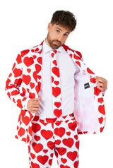 A man is dressed in the Love - Heart suit from Suitmeister, featuring a white suit adorned with large red hearts. He holds the jacket open to showcase its interior design. Complementing the ensemble is a matching tie with the same heart pattern, worn over a crisp white shirt. The background is a simple white.