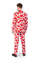 A man is standing facing away wearing the "Love - Heart suit" by Suitmeister. The predominantly white suit features bold red heart patterns. He has paired it with black shoes, and the background is plain white.