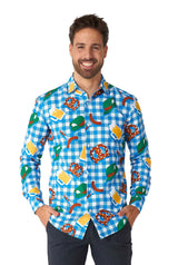 A man wearing the October Check Blue - Octoberfest shirt by Suitmeister, featuring playful patterns of beer mugs, pretzels, and sausages on a blue and white checkered design, smiles with his hands in his pockets against a white background.