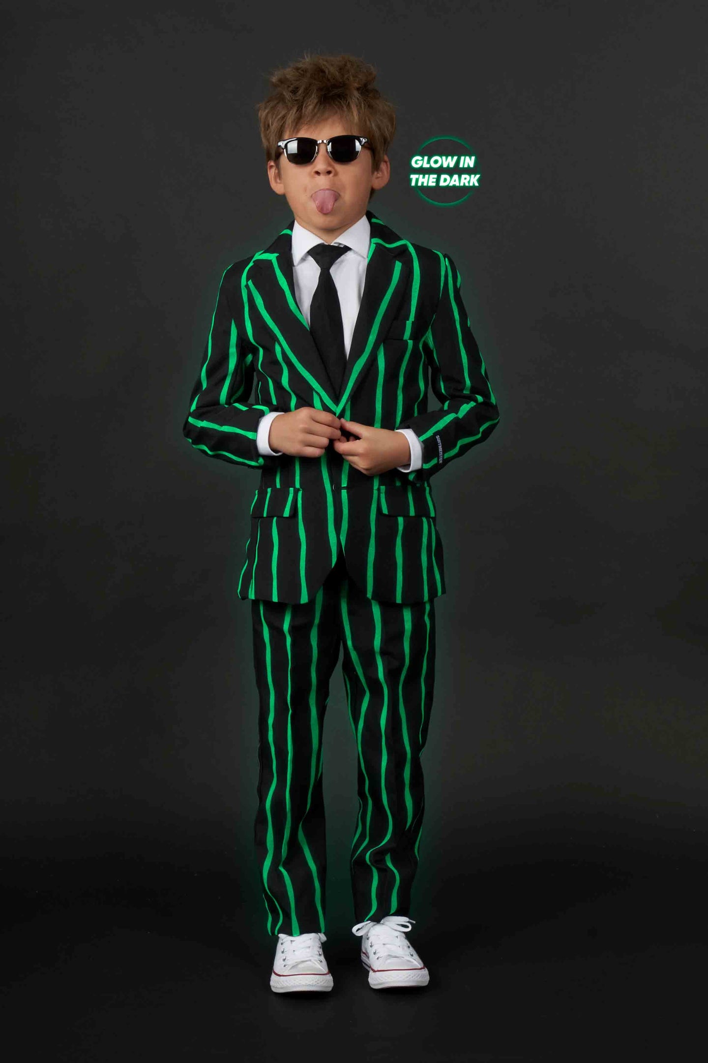 A child sticks out their tongue while wearing an oversized pinstripe black suit and tie from Suitmeister. They have sunglasses and white sneakers, standing against a dark background.