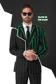 A man in an Oversized Pinstripe Black suit by Suitmeister, featuring fluorescent green stripes on one side, paired with sunglasses and a black tie, stands against a contrasting split black and white background. The phrase "Glow in the Dark" is displayed in the upper right corner.