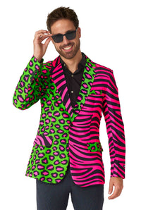 A bearded individual wearing the Suitmeister Party Animal Neon Party blazer, featuring neon pink zebra stripes and green leopard spot patterns, smiles at the camera while holding their sunglasses with one hand.