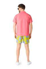 A man stands facing away, dressed in a Patrick costume by Suitmeister, consisting of a pink short-sleeve shirt, yellow and blue patterned shorts, and white sneakers. He accessorizes with sunglasses against a plain white background.