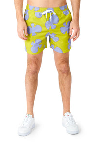 A person is dressed in the Patrick costume by Suitmeister, featuring lime green swim shorts adorned with light purple puzzle piece patterns, standing against a white background and wearing white sneakers.