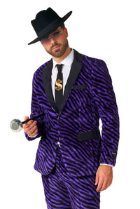 A person is dressed in the Suitmeister "Pimp Faux Fur Purple" ensemble, including a purple zebra-striped suit with matching pants. They accessorize with a black hat and sunglasses while holding a faux diamond-topped cane. The outfit includes a tie adorned with a gold dollar sign, and the person's expression is neutral, set against a white background.