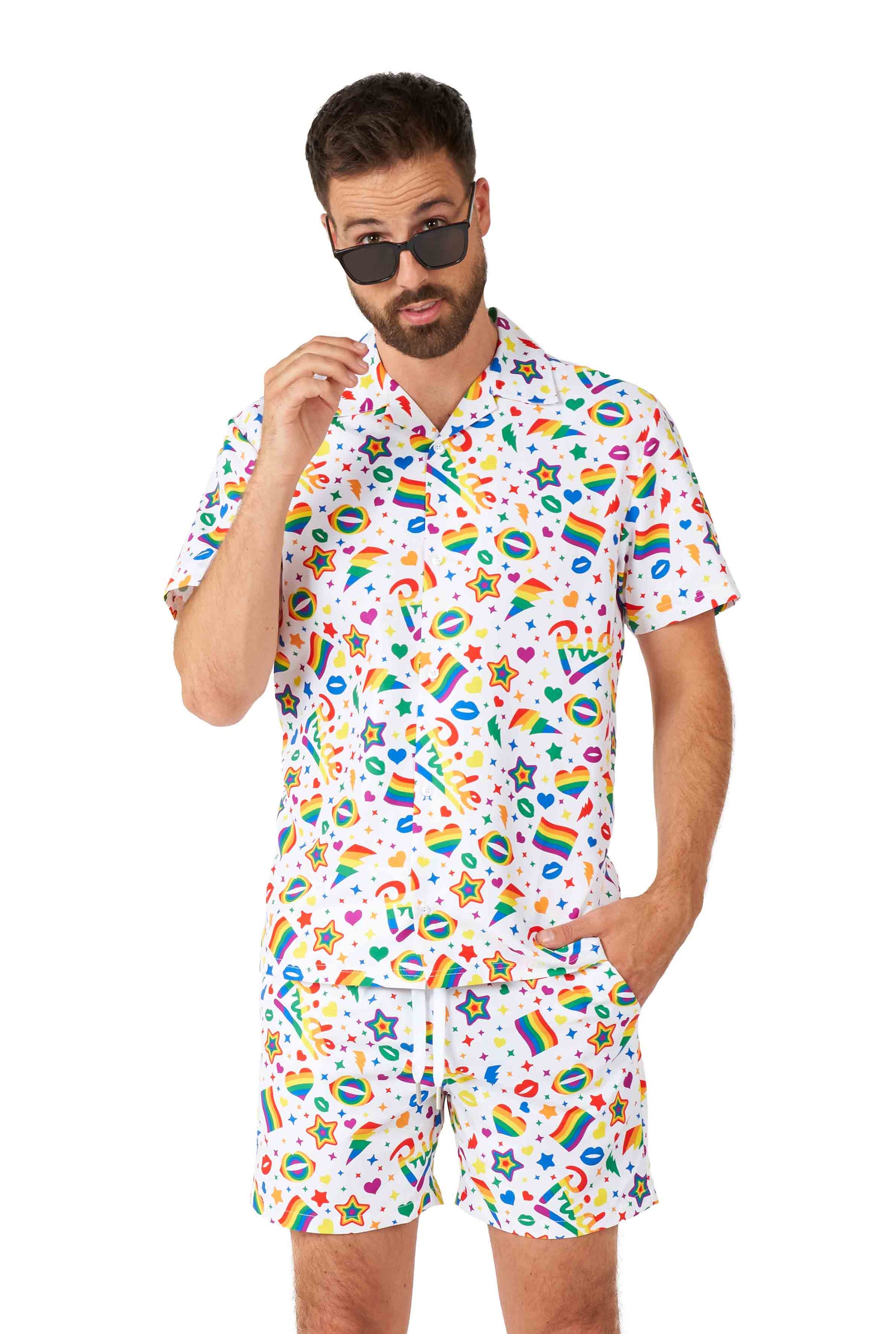 A man wearing the Pride Icons White - Pride outfit by Suitmeister, featuring sunglasses and a colorful matching shirt and shorts adorned with playful prints of hearts, rainbows, stars, and squiggles, looks at the camera with one hand in his pocket.