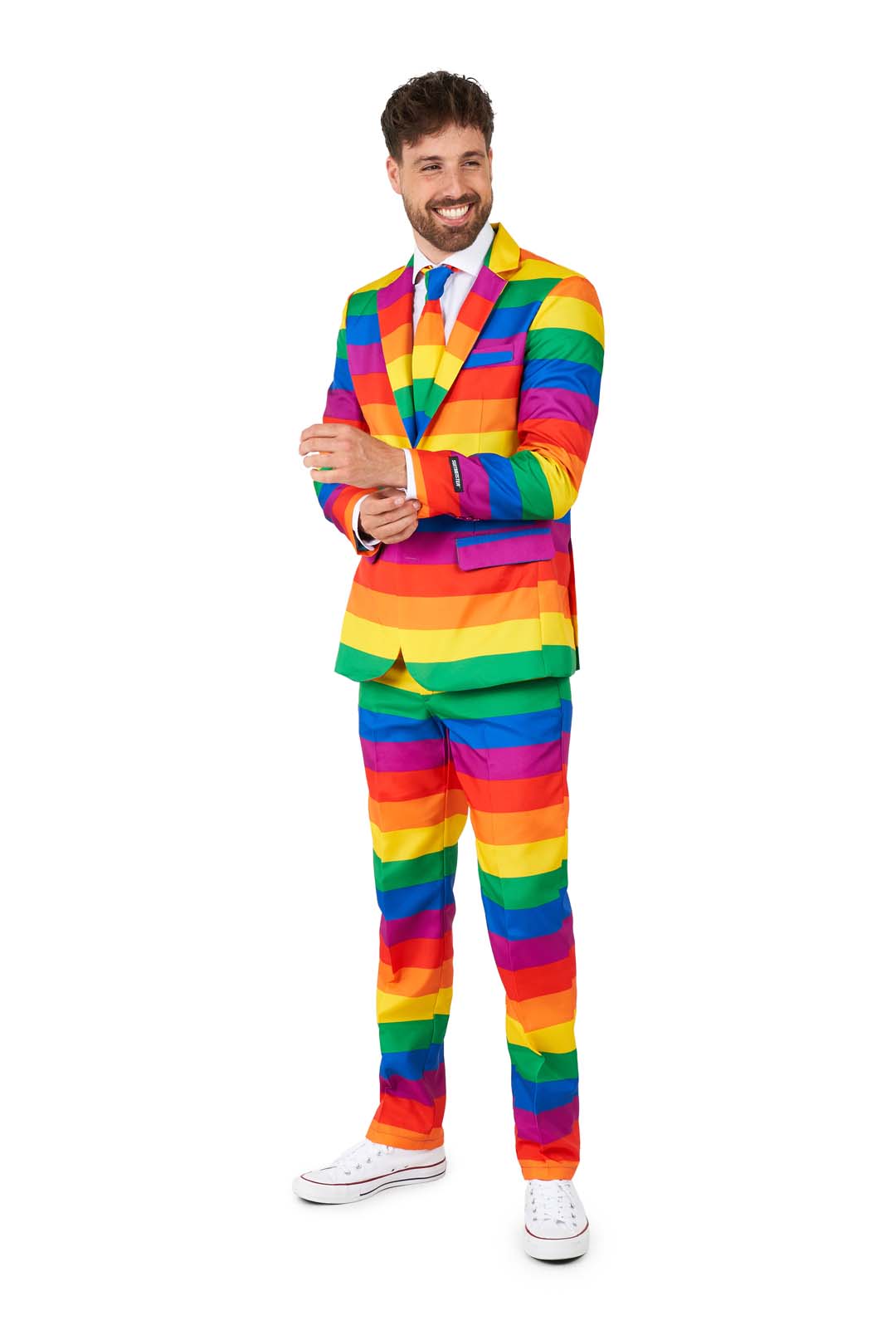 A smiling person wearing the Suitmeister Rainbow suit stands against a white background. The ensemble includes a colorful rainbow-striped jacket and matching pants, paired with white sneakers. They are adjusting their sleeve with one hand.